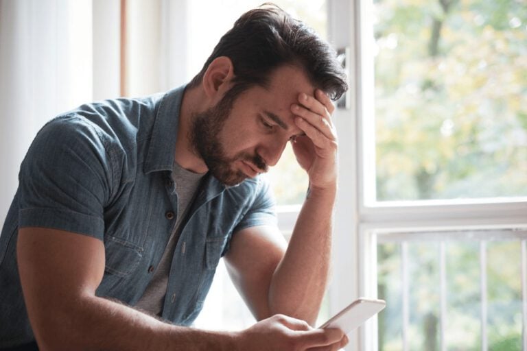 A man looking at his phone while sitting in front of a window.