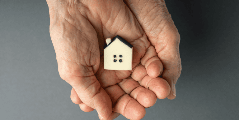 Two hands gently cradling a small, symbolic white house with a black roof, conveying the concept of home care, ownership, protection, or real estate.