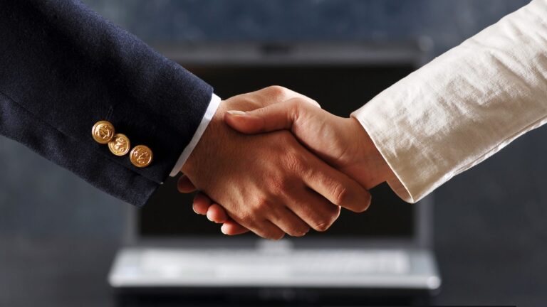 שני אנשי מקצוע בלבוש רשמי לוחצים ידיים על מחשב נייד, מסמנים הסכם, שיתוף פעולה או חתימה של עסקה.