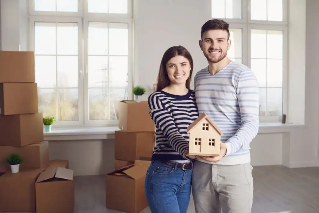 זוג חייכן עומד מול ערימת קופסאות מעבר בחדר מואר. שניהם אוחזים בדגם בית עץ קטן. האישה לובשת