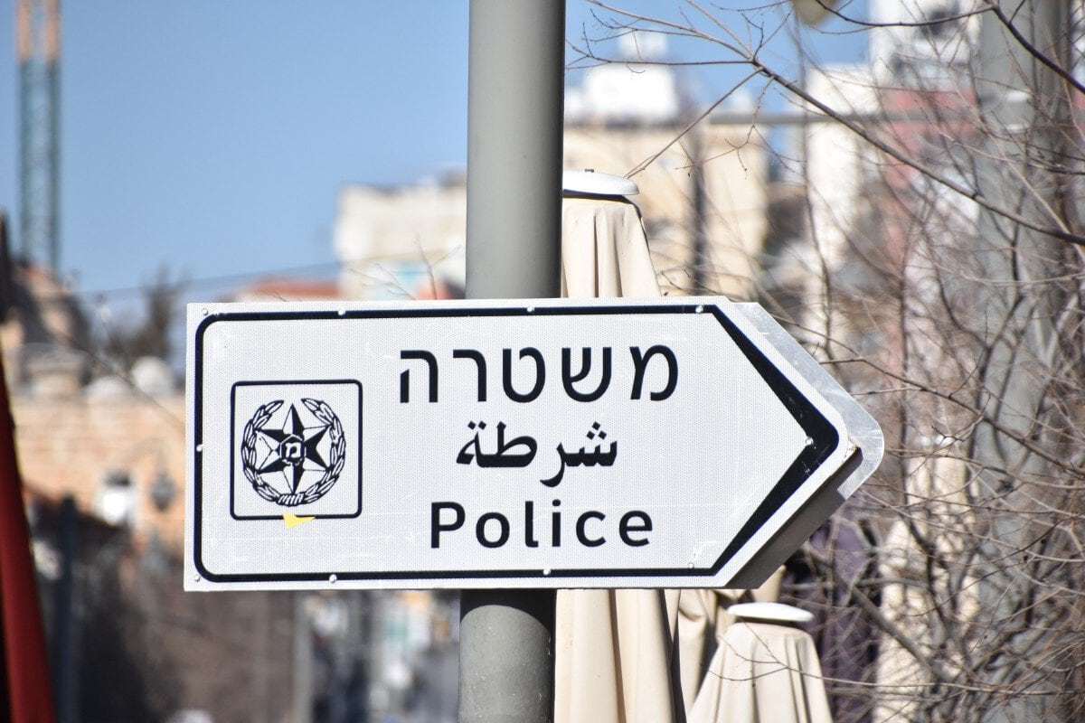מור ושות׳ חברת עורכי דין ונוטריון שלט רחוב המותקן על עמוד מצביע על תחנת משטרה. השלט כולל טקסט בעברית, ערבית ואנגלית, האומר "משטרה", יחד עם סמל. ברקע נראה סביבה עירונית מטושטשת עם מבנים ועצים.