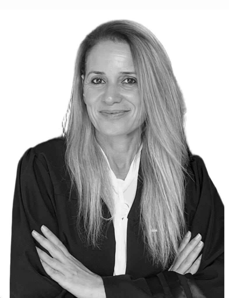 אישה עם שיער ארוך וישר, לבושה בגלימה שחורה וחולצה לבנה, עומדת עם זרועות שלובות ומחייכת למצלמה על רקע לבן חלק.