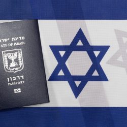 דרכון ישראלי שחור מונח על משטח עם דגל ישראל. לדגל יש פסים כחולים אופקיים למעלה ולמטה עם מגן דוד כחול במרכז. הדרכון מציג את סמל ישראל וטקסט בעברית ובאנגלית.