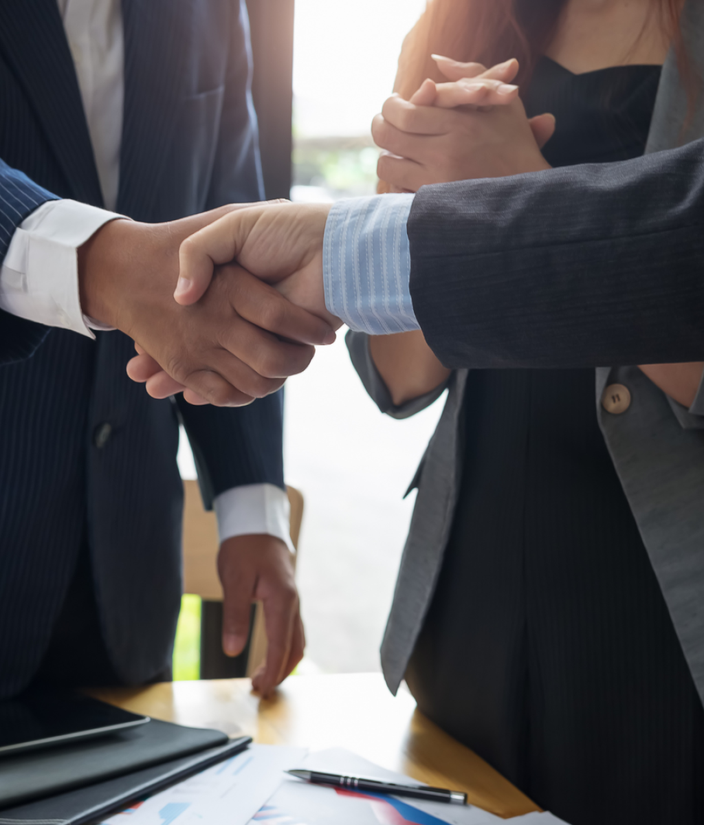 שלושה אנשים בלבוש עסקי עומדים ולוחצים ידיים בסביבה ארגונית. שני האנשים בחזית לוחצים ידיים, האחד לובש חליפה כחולה והשני בחליפה אפורה. אדם שלישי בבלייזר אפור עומד בסמוך עם ידיים שלובות.